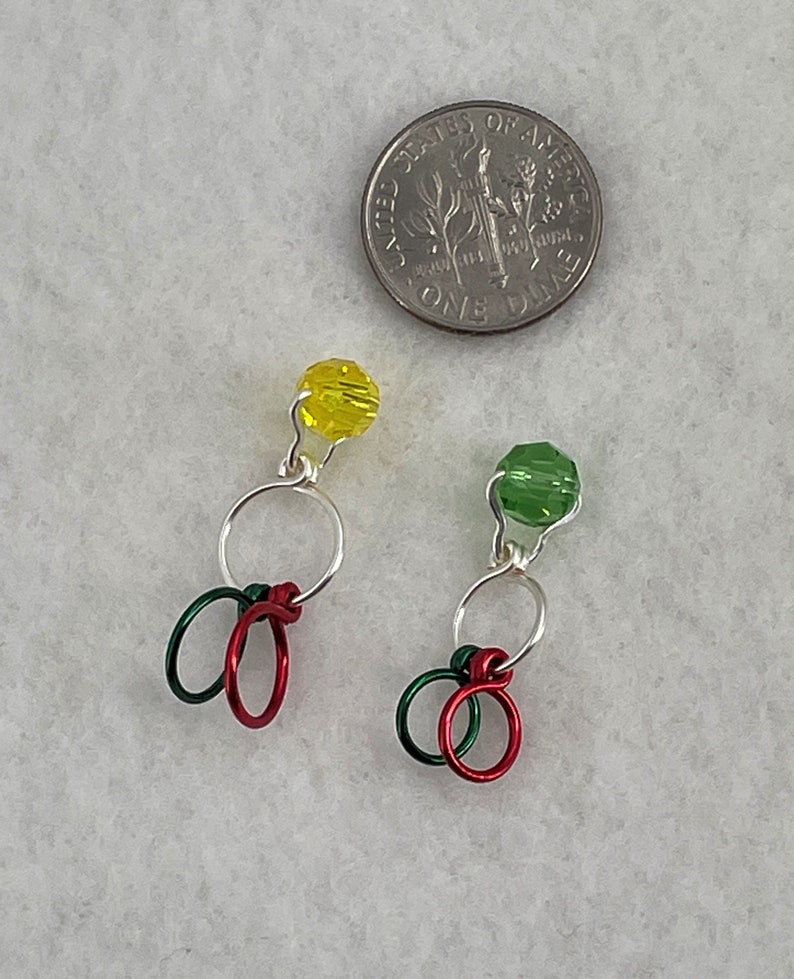 May include: Two silver wire stitch markers with red and green loops and a yellow and green glass bead at the top.