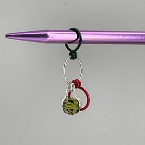 K&ouml;nnte beinhalten: Eine gr&uuml;ne Glasperle mit einer silbernen und roten Drahtschlaufe. Die Schlaufe ist an einer lila Stricknadel befestigt.