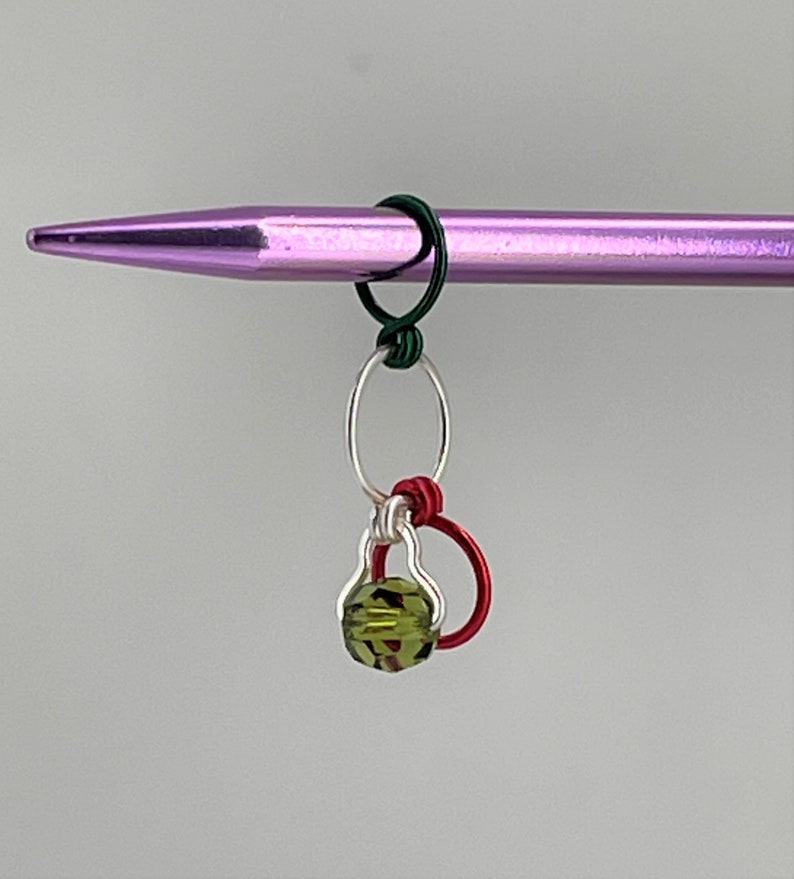 K&ouml;nnte beinhalten: Eine gr&uuml;ne Glasperle mit einer silbernen und roten Drahtschlaufe. Die Schlaufe ist an einer lila Stricknadel befestigt.