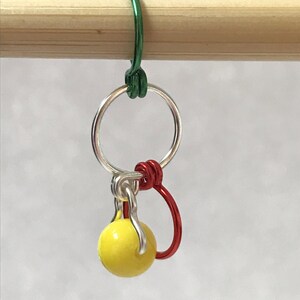 May include: A yellow bead stitch marker with a silver ring and red and green wire loops.