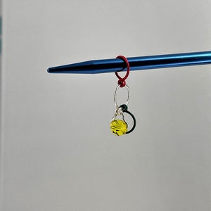 May include: A yellow glass bead charm with a silver ring and a red and green wire loop attached to a blue knitting needle.