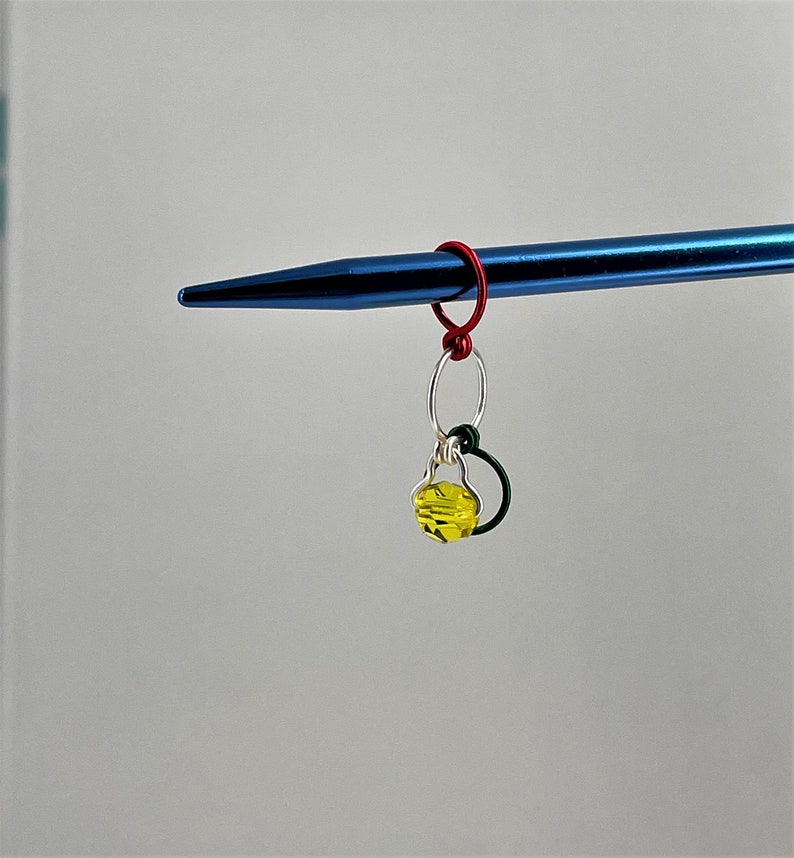 May include: A yellow glass bead charm with a silver ring and a red and green wire loop attached to a blue knitting needle.