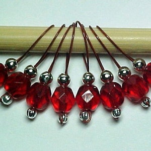 May include: Set of eight red glass bead stitch markers with silver metal caps, attached to brown cords, displayed on a wooden dowel.