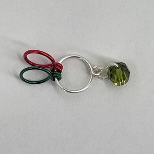 K&ouml;nnte beinhalten: Ein silberner Ring, um den ein gr&uuml;ner und roter Draht gewickelt ist. Ein gr&uuml;ner Kristall h&auml;ngt an dem Ring.