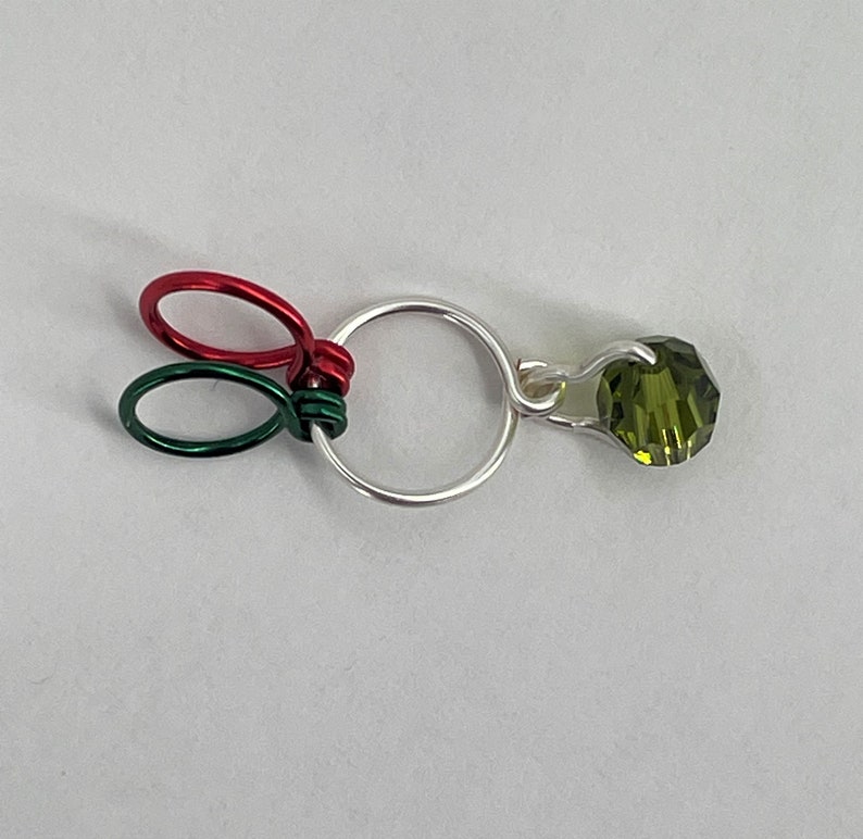 K&ouml;nnte beinhalten: Ein silberner Ring, um den ein gr&uuml;ner und roter Draht gewickelt ist. Ein gr&uuml;ner Kristall h&auml;ngt an dem Ring.