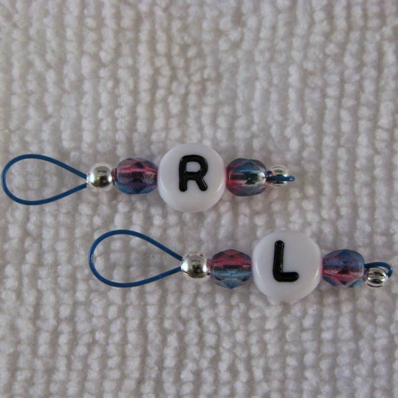 May include: Two white bead charms with blue and pink accents. The beads have the letters "R" and "L" on them. Each bead has a blue loop attached.