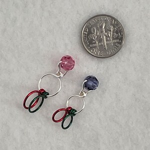 May include: Two pairs of colorful stitch markers with silver rings and beads. One pair has pink beads and red and green wire. The other pair has purple beads and green wire.
