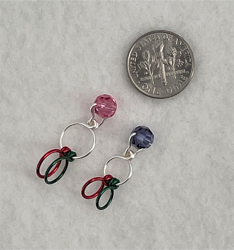 May include: Two pairs of colorful stitch markers with silver rings and beads. One pair has pink beads and red and green wire. The other pair has purple beads and green wire.