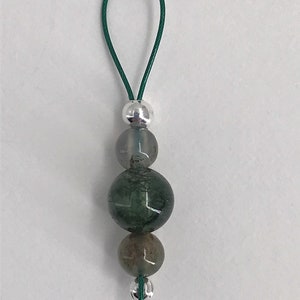 Puede incluir: Un amuleto de cuentas de &aacute;gata verde y gris con una cuenta de plata y un cord&oacute;n verde.