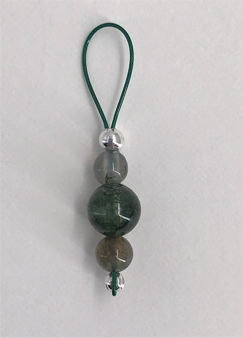 Puede incluir: Un amuleto de cuentas de &aacute;gata verde y gris con una cuenta de plata y un cord&oacute;n verde.