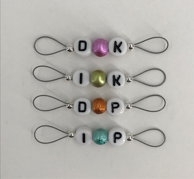 May include: Four sets of white letter beads with silver loops. The beads spell out "DIK", "IK", "DP", and "IP". The beads are decorated with different colored accents, including pink, green, orange, and turquoise.