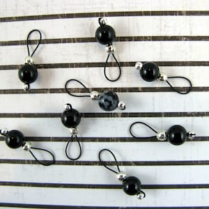 May include: Eight black and white stitch markers with silver rings. The stitch markers are made of glass and have a variety of patterns.