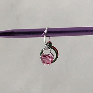 May include: A pink crystal bead charm with a silver wire loop and green and red wire accents. The charm is attached to a purple knitting needle.