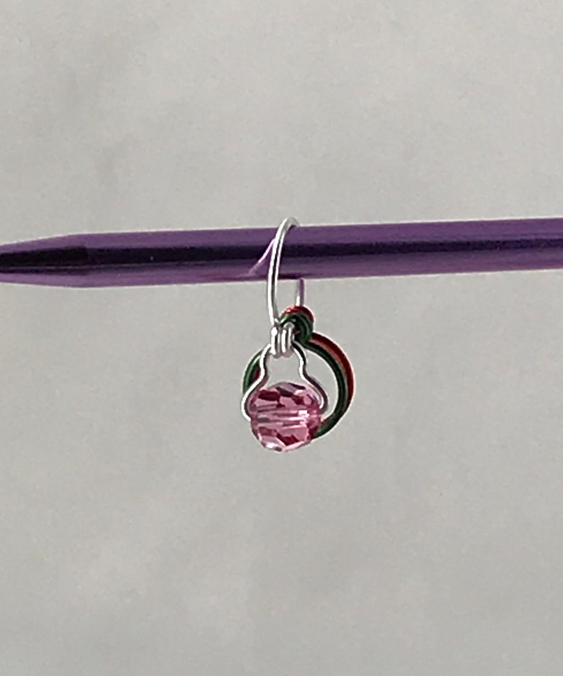 May include: A pink crystal bead charm with a silver wire loop and green and red wire accents. The charm is attached to a purple knitting needle.