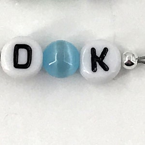 May include: A white beaded keychain with the letters "D" and "K" separated by a light blue bead. The keychain has a silver loop on each end.