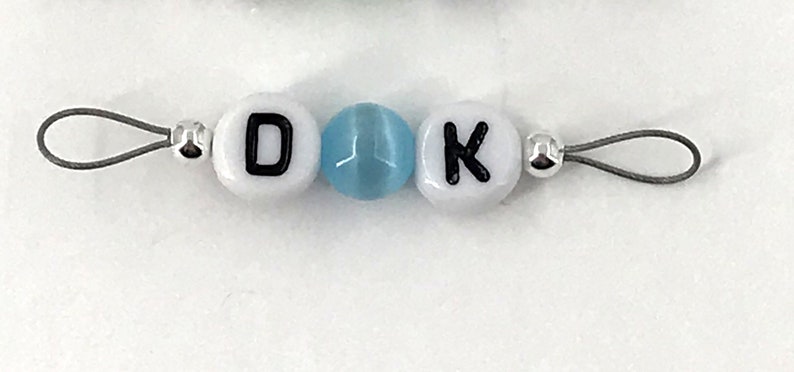 May include: A white beaded keychain with the letters "D" and "K" separated by a light blue bead. The keychain has a silver loop on each end.