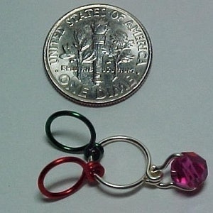 Pu&ograve; includere: Un primo piano di un ciondolo d'argento con un cristallo rosa, tre anelli metallici, uno verde e due rossi.