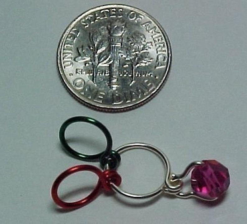 Pu&ograve; includere: Un primo piano di un ciondolo d'argento con un cristallo rosa, tre anelli metallici, uno verde e due rossi.