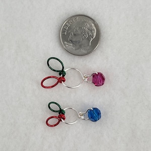 May include: Two silver wire rings with a red and green wire loop attached to each. A pink crystal is attached to one ring and a blue crystal is attached to the other ring.