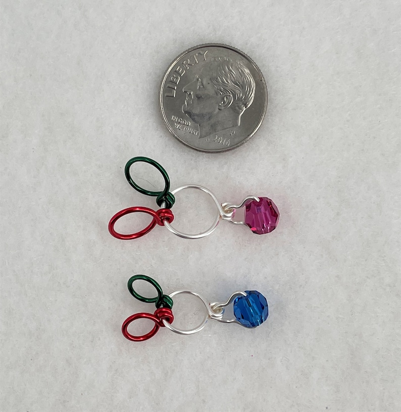 Pu&ograve; includere: Due anelli di filo d'argento con un anello di filo rosso e verde attaccato a ciascuno. Un cristallo rosa &egrave; attaccato a un anello e un cristallo blu &egrave; attaccato all'altro anello.