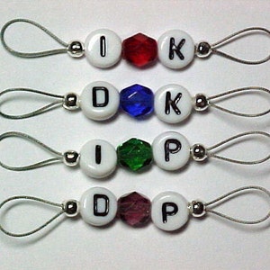 May include: Four white bead charms with black letters spelling out "I D K P" and four colored glass beads. Each bead charm is attached to a silver loop.