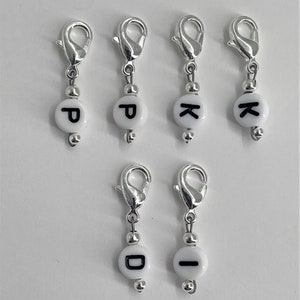 May include: Six silver stitch markers with white beads and black letter charms. The letters are P, K, D, and I.