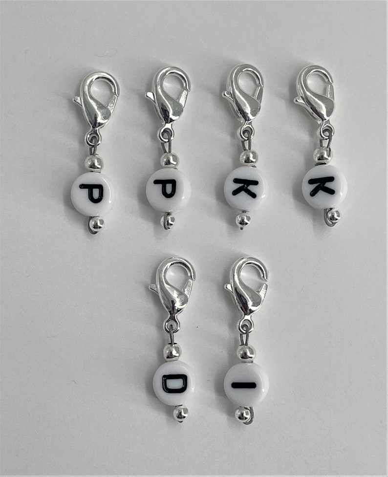 Puede incluir: Seis marcadores de punto de plata con cuentas blancas y abalorios de letras negras. Las letras son P, K, D e I.