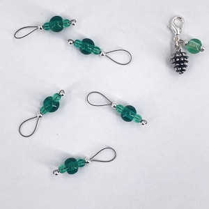 May include: Five stitch markers featuring dark green glass beads and silver-toned metal loops, alongside one silver-toned metal lobster clasp charm featuring a small pinecone pendant and a single matching green glass bead.