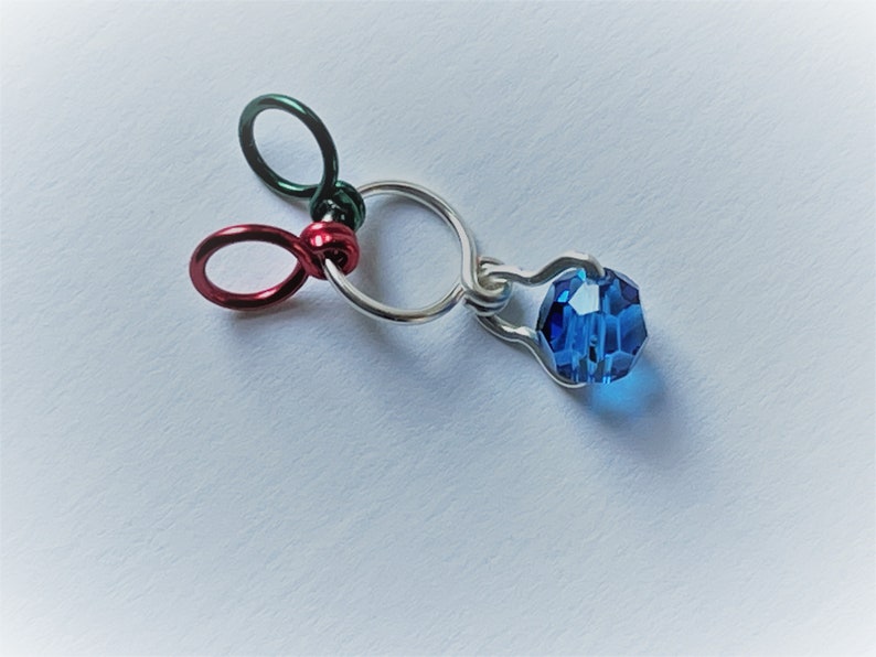 K&ouml;nnte beinhalten: Ein silberner Anh&auml;nger mit einer blauen Kristallperle, befestigt an drei kleinen Ringen in Rot, Gr&uuml;n und Silber.
