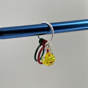May include: A silver knitting stitch marker with three colorful rings and a yellow faceted bead. The rings are red, green, and silver.