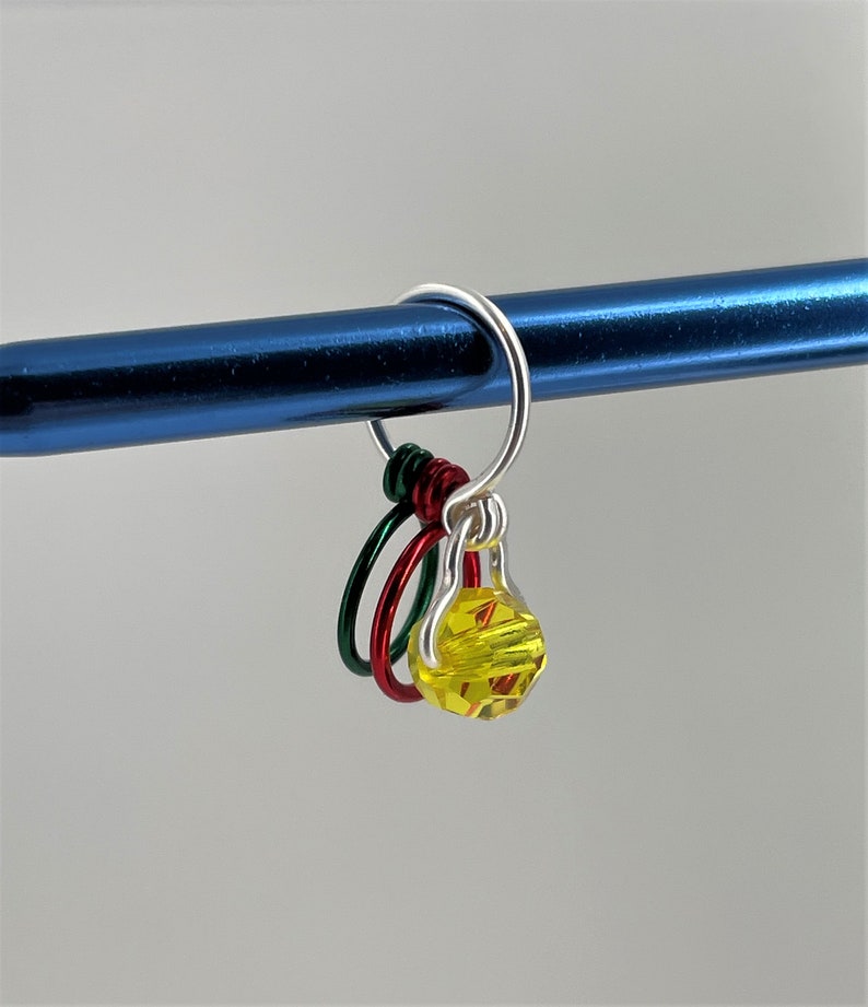 May include: A silver knitting stitch marker with three colorful rings and a yellow faceted bead. The rings are red, green, and silver.