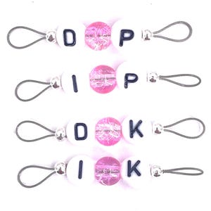 May include: Four zipper pulls featuring white round beads with black letters spelling DP, IP, DK, and IK. Each pull includes a central translucent pink crackle bead, silver-toned metal spacers, and a flexible gray wire loop for attachment to zippers.