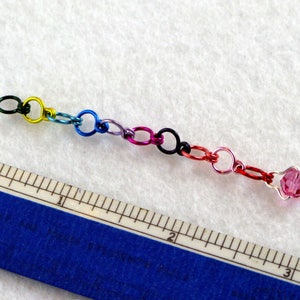 May include: A colorful chain made of small metal rings in shades of green, yellow, blue, purple, pink, black, and red, with a pink crystal bead at the end. The chain is about 2 inches long and is measured against a ruler.