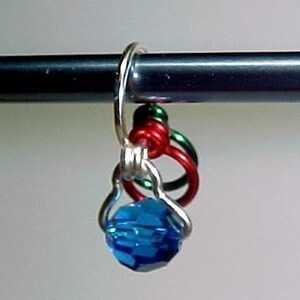 Pu&ograve; includere: Un perno di cristallo blu con un anello di filo d'argento attaccato a tre anelli di filo colorati. Gli anelli sono rosso, verde e blu.
