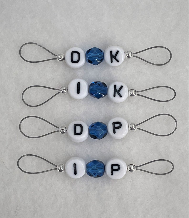 Może przedstawiać: Cztery zestawy białych koralik&oacute;w z literami i niebieskimi koralikami kryształowymi na srebrnym sznurku. Litery tworzą słowa "DOK", "IK", "DOP" i "IP".