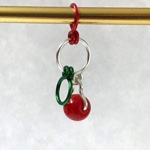 May include: A red and green charm with a red cherry bead, a silver ring, and a green ring. The charm is attached to a gold metal rod.