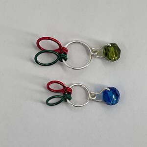 May include: Two silver wire rings with green and red wire loops and a crystal bead attached to each ring. The top bead is green and the bottom bead is blue.