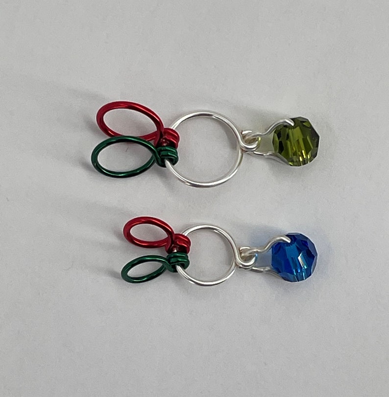 K&ouml;nnte beinhalten: Zwei silberne Drahtringe mit gr&uuml;nen und roten Drahtschlaufen und einer Kristallperle, die an jedem Ring befestigt ist. Die obere Perle ist gr&uuml;n und die untere Perle ist blau.