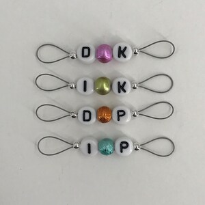 May include: Four sets of white letter beads with black lettering, strung on silver wire loops. Each set spells out a different name: DIK, IKK, DOP, and IPP. The beads are interspersed with colorful, metallic beads.