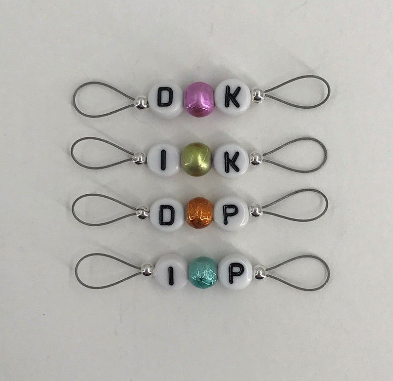May include: Four sets of white letter beads with black lettering, strung on silver wire loops. Each set spells out a different name: DIK, IKK, DOP, and IPP. The beads are interspersed with colorful, metallic beads.
