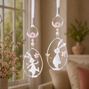 Könnte beinhalten: Zwei Osterei-Ornamente mit weißen Hasen-Silhouetten und rosa Blumenakzenten. Jedes Ornament hängt an einem weißen Band und hat einen dekorativen Ring mit Perlen und Blumen. Der Hintergrund umfasst ein Fenster und eine Blumenvase.