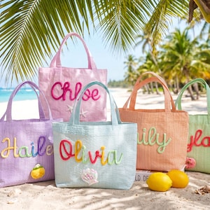 Könnte beinhalten: Fünf farbenfrohe Seersucker-Tragetaschen mit gestickten Namen: Chloe, Hailee, Olivia, Lily und Isla. Jede Tasche hat ein anderes Farbschema und dekorative Akzente wie Muscheln und Seesterne. Die Taschen stehen auf einem Sandstrand mit Palmen.