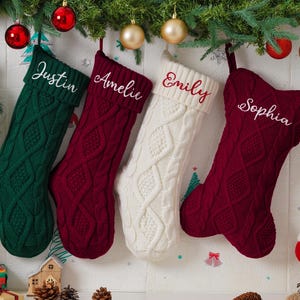 May include: Four cable-knit Christmas stockings in green, burgundy, and white hang from a garland with ornaments. Each stocking is personalized with a name embroidered in a contrasting color: Justin, Amelie, Emily, and Sophia.