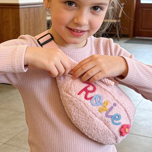 May include: A pale pink fanny pack with the name "Rosie" embroidered in colourful thread. The pack has a soft, textured appearance and a small flower appliqué. The adjustable strap is black. The fanny pack is designed for children.