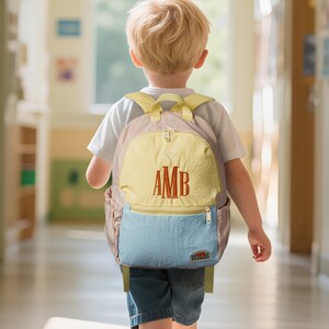 May include: A multi-coloured backpack with the embroidered initials "AMB" in a red colour. The backpack has a yellow front panel, a light blue bottom pocket, and a light pink body. The backpack is being worn by a child.