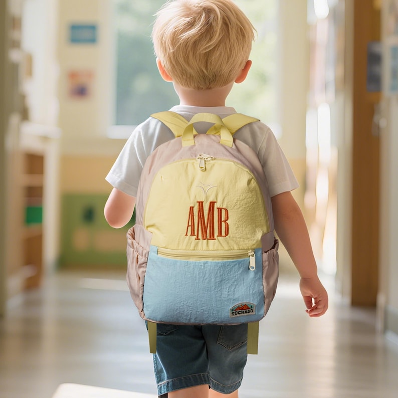 May include: A multi-coloured backpack with the embroidered initials "AMB" in a red colour. The backpack has a yellow front panel, a light blue bottom pocket, and a light pink body. The backpack is being worn by a child.