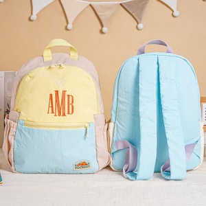 May include: Two colourful backpacks. One is light blue, yellow, and pink with the embroidered letters "AMB". The other is light blue with purple accents. Both have adjustable straps and side pockets. The backpacks are on a white surface.
