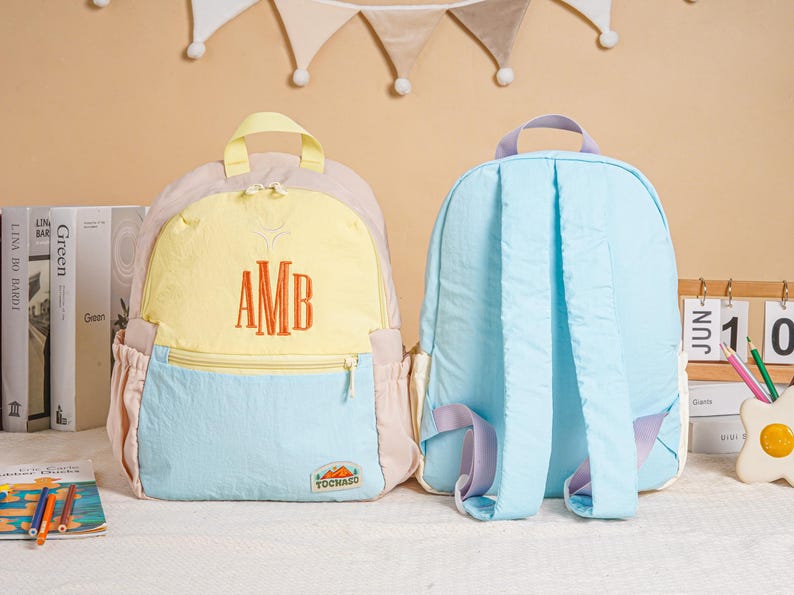May include: Two colourful backpacks. One is light blue, yellow, and pink with the embroidered letters "AMB". The other is light blue with purple accents. Both have adjustable straps and side pockets. The backpacks are on a white surface.