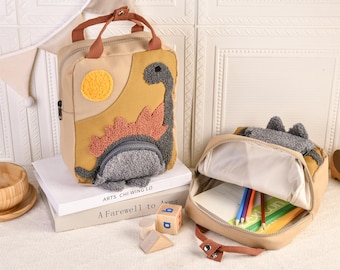 May include: Tan dinosaur-themed backpack with a yellow sun and a gray dinosaur design. The backpack has a small front pocket and a top handle. A second open backpack with pencils and paper is next to it. The backpack is sitting on a stack of books.