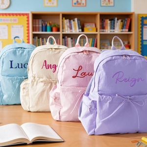 May include: Four pastel-coloured backpacks, each embroidered with a name: "Luc", "Ang", "Lau", and "Reign". The colours are light blue, cream, pink, and lavender. An open book and pencils are on the wooden table in front of the backpacks.
